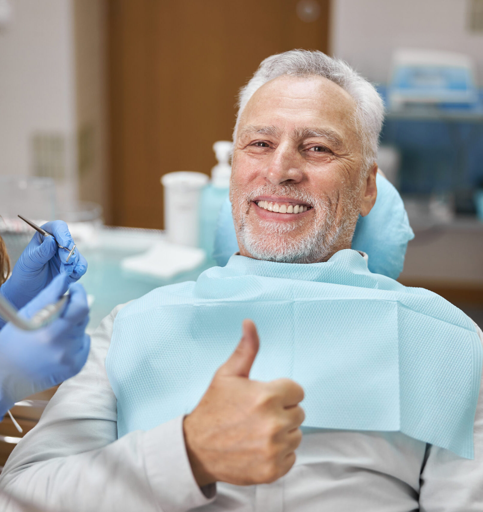 Mirthful senior citizen smiling and showing thumbs up at dental appointment with his dentist and looking at the camera